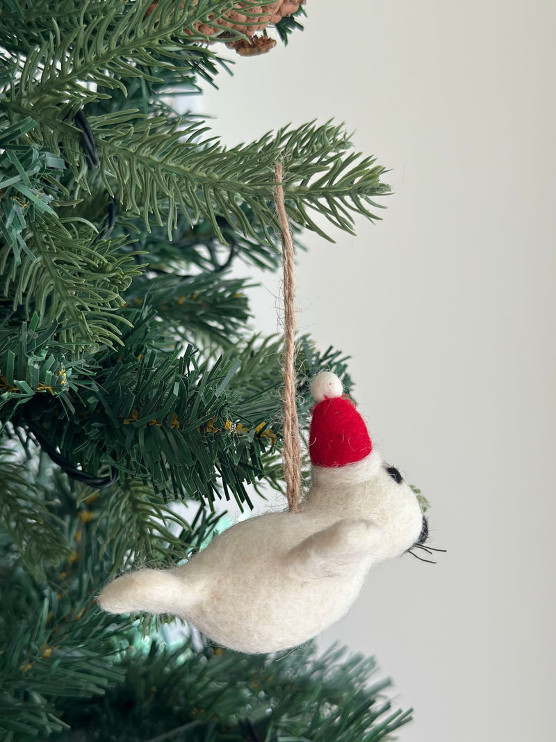 A white felt mini seal ornament, wearing a red Christmas hat with a white pompom, hangs from a lush green Christmas tree branch by a jute string. The charming handmade felt ornament features tiny black eyes and whiskers, adding a cheerful touch to holiday decor.