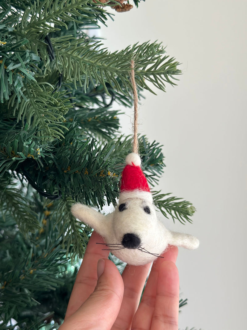 A felt mini seal Christmas ornament, wearing a red Christmas hat, is held by a female hand against a festive Christmas tree. This charming white felt seal ornament features black felt dots for eyes and nose, along with delicate whiskers. A fluffy white pompom adorns the red Christmas hat, which has a jute string attached for hanging.