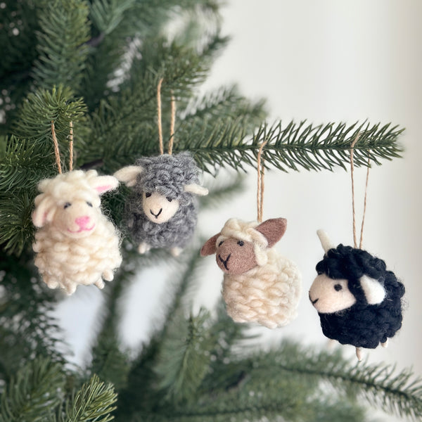 Four wool felt mini sheep ornaments are displayed on a lush green Christmas tree. These handcrafted, needle felted sheep ornaments come in white, gray, beige, and black. Each 3D sheep features small ears and a stitched mouth, suspended by a jute string, enhancing a warm holiday feel.