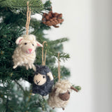 Joyful felt mini sheep ornaments, handmade from organic wool, adorn the green branches of a Christmas tree. These charming needle felted ornaments, suspended by jute strings, appear in white, black, and cream, each with a brown face, stitched smile, and small felted ears. Their fluffy texture mimics real wool, creating a cozy and festive atmosphere.
