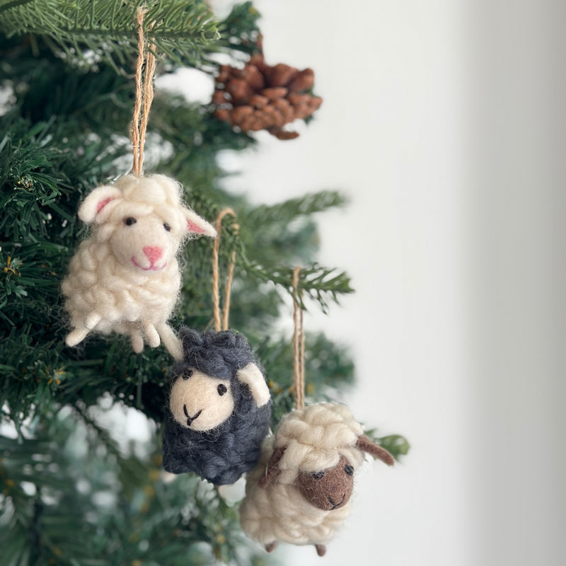 Joyful felt mini sheep ornaments, handmade from organic wool, adorn the green branches of a Christmas tree. These charming needle felted ornaments, suspended by jute strings, appear in white, black, and cream, each with a brown face, stitched smile, and small felted ears. Their fluffy texture mimics real wool, creating a cozy and festive atmosphere.