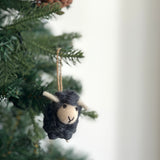 A needle felted mini sheep ornament hangs on a green Christmas tree branch. This 3D sheep ornament features a black body, a beige face with hand-stitched eyes and mouth, and small white horns. A natural jute string is attached for hanging.