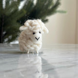 A cute felt mini sheep ornament, ideal as a table decoration, is displayed on a glossy marble surface before a blurred green tree. This charming handmade ornament, crafted from fluffy off-white wool, features a rounded body, small black eyes, and a stitched smiling mouth. It wears a colorful scarf and has delicate thin legs, with a hemp string loop for hanging.