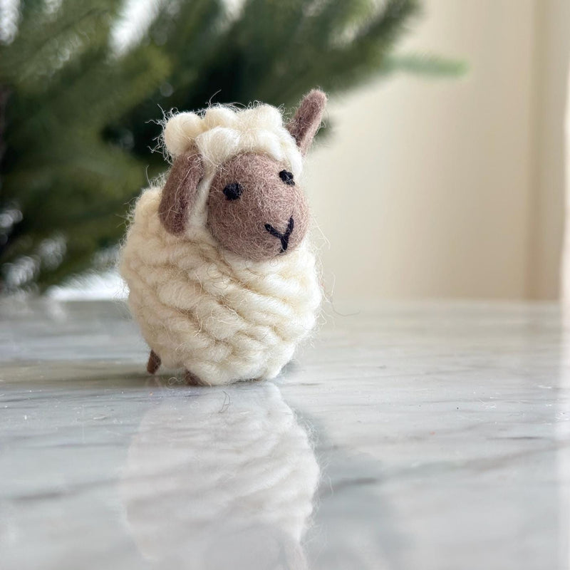 A charming mini wool felt sheep Christmas ornament rests on a reflective marble surface, with a blurred green backdrop. This needle felted sheep features a round, fluffy white body with a light brown face and ears, tiny black stitched eyes and mouth, and a scarf.