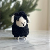 A charming wool felt mini sheep ornament, ideal as a table decoration, rests on a white marble surface. This handmade ornament features a fluffy black wool body, a white face with a black stitched 'Y' for its mouth, and small white legs. It has white wool ears and wears a cozy scarf, all set against a soft-focused backdrop of a green, leafy tree.