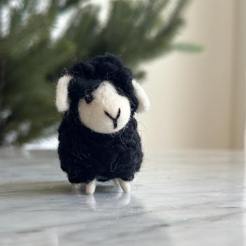 A charming wool felt mini sheep ornament, ideal as a table decoration, rests on a white marble surface. This handmade ornament features a fluffy black wool body, a white face with a black stitched 'Y' for its mouth, and small white legs. It has white wool ears and wears a cozy scarf, all set against a soft-focused backdrop of a green, leafy tree.