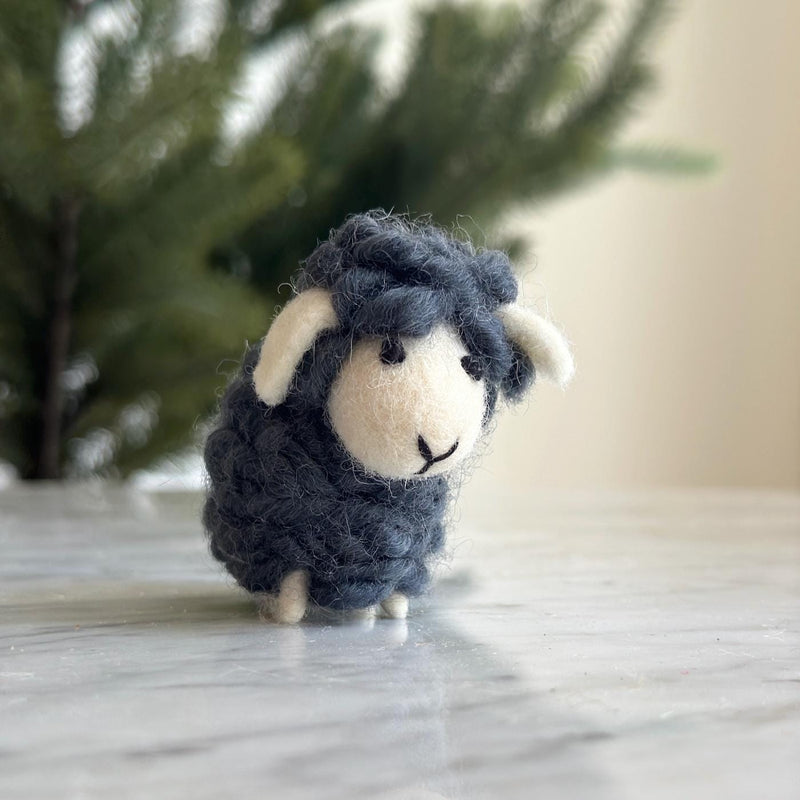 Felt mini sheep decorations are presented, hanging on a blurred Christmas tree, suggesting a cozy holiday atmosphere. This charming wool felt mini sheep ornament features a white face with stitched black eyes and mouth, white ears, and a soft gray, fuzzy body adorned with a scarf.