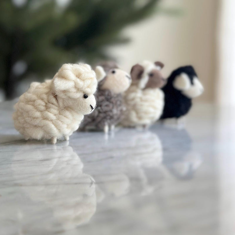 Multiple wool felt mini sheep ornaments are displayed in a neat row on a reflective surface, set against a blurred green, festive background. These handmade felt sheep come in white, gray, tan, and black variations. Each needle-felted sheep features a round body, fuzzy fur, and a small scarf, showcasing their cozy, handcrafted charm.