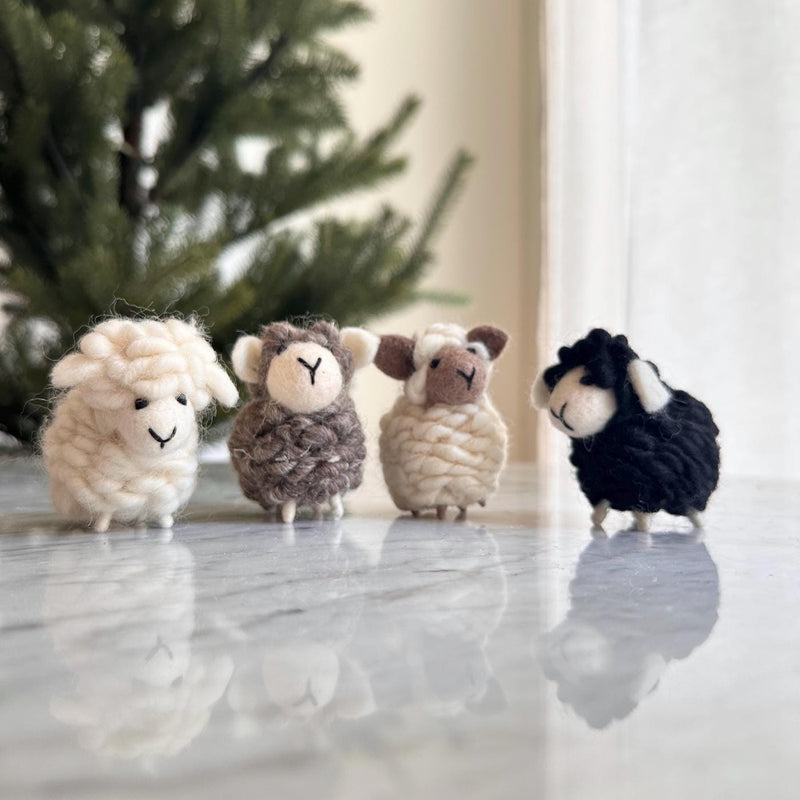 Four unique wool felt sheep ornaments, suitable as charming felt mini sheep table decorations, stand in a row on a reflective marble surface. A blurred Christmas tree and a window with sheer white curtains form the serene background. These handmade felt sheep feature a fluffy texture and unique colors: one white, one brown, one cream and brown, and one black. Each sheep has small upright ears, tiny legs, an embroidered 'Y' nose, and a curved stitch mouth.