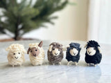 Handmade wool felt mini sheep ornaments are displayed in a row on a white marble surface, with a hint of a pine tree in the bright background. These cozy sheep ornaments feature a round body with fuzzy fur, tiny ears, a stitched smile, and a colorful scarf. A hemp string loop is visible for hanging. They come in white, light beige, brown, dark blue, and black, with one showing brown ears and face.