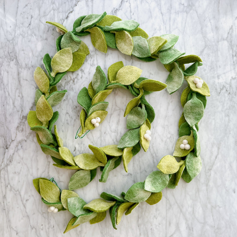 A handmade Felt Mistletoe Garland is presented in a spiral on a white marble surface. This whimsical wool felt garland features leaves in varying shades of green, interspersed with clusters of small, white wool felt berries, all suspended on a natural jute thread, highlighting its festive and natural appeal.