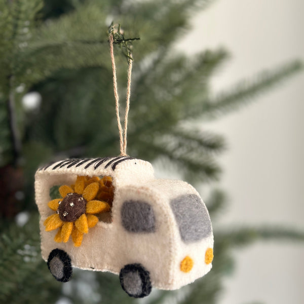 The Felt Mobile Flower Shop Christmas ornament is displayed on a Christmas tree. This charming unique felt ornament features a white felt truck with black stripes, overflowing with bright yellow sunflowers that have brown, beaded centers. The truck includes gray windows, yellow lights, black wheels, and a jute string for easy hanging.