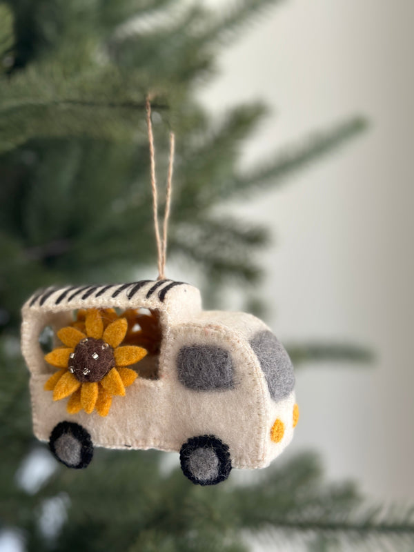 A handcrafted felt mobile flower shop ornament is displayed hanging on a Christmas tree. This felt ornament for Christmas features a white truck brimming with yellow sunflowers. The truck's top is striped in black and white, with black tires and grey centers. A jute string allows for easy hanging, adding a whimsical and joyful touch to the festive decor.