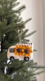 A unique Felt Mobile Flower Shop Christmas tree ornament is shown hanging on a green Christmas tree. This handcrafted felt ornament features a white truck with gray windows, wheels, and a black and white striped roof rack. Two bright yellow sunflowers with beaded brown centers adorn the truck's side. It hangs from a jute string, blending with the tree's green needles.
