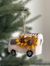 This unique felt flower shop truck ornament hangs from a tree branch, set against blurred evergreen foliage and a light gray wall. The white felt truck, adorned with vibrant yellow sunflowers, features gray accents, black stripes on top, and black wheels. Each felt sunflower boasts a brown center dotted with tiny silver beads. A jute string is attached for easy hanging, making it a delightful Christmas ornament.