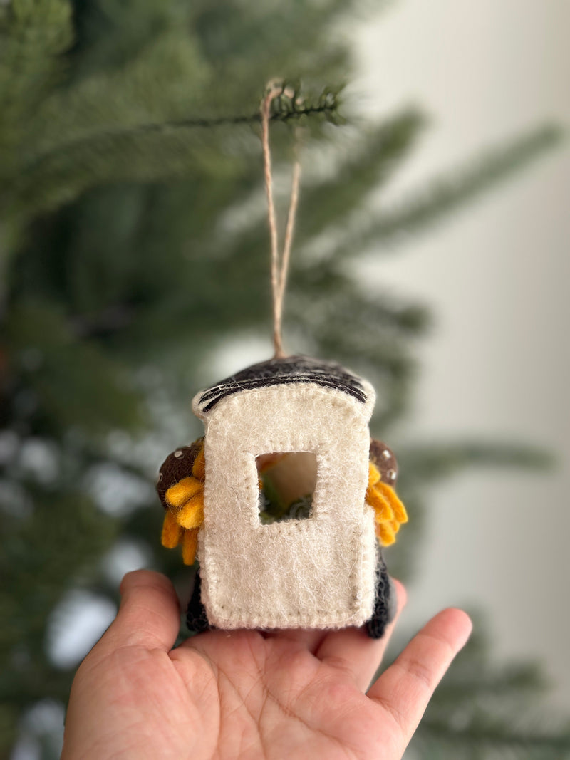 This charming Felt Mobile Flower Shop Christmas ornament is held in a hand, set against a softly blurred Christmas tree backdrop. The white felt truck, featuring a gray roof and dark gray base, is brimming with vibrant yellow sunflowers that have brown centers and yellow petals. A natural jute string is attached for easy hanging.