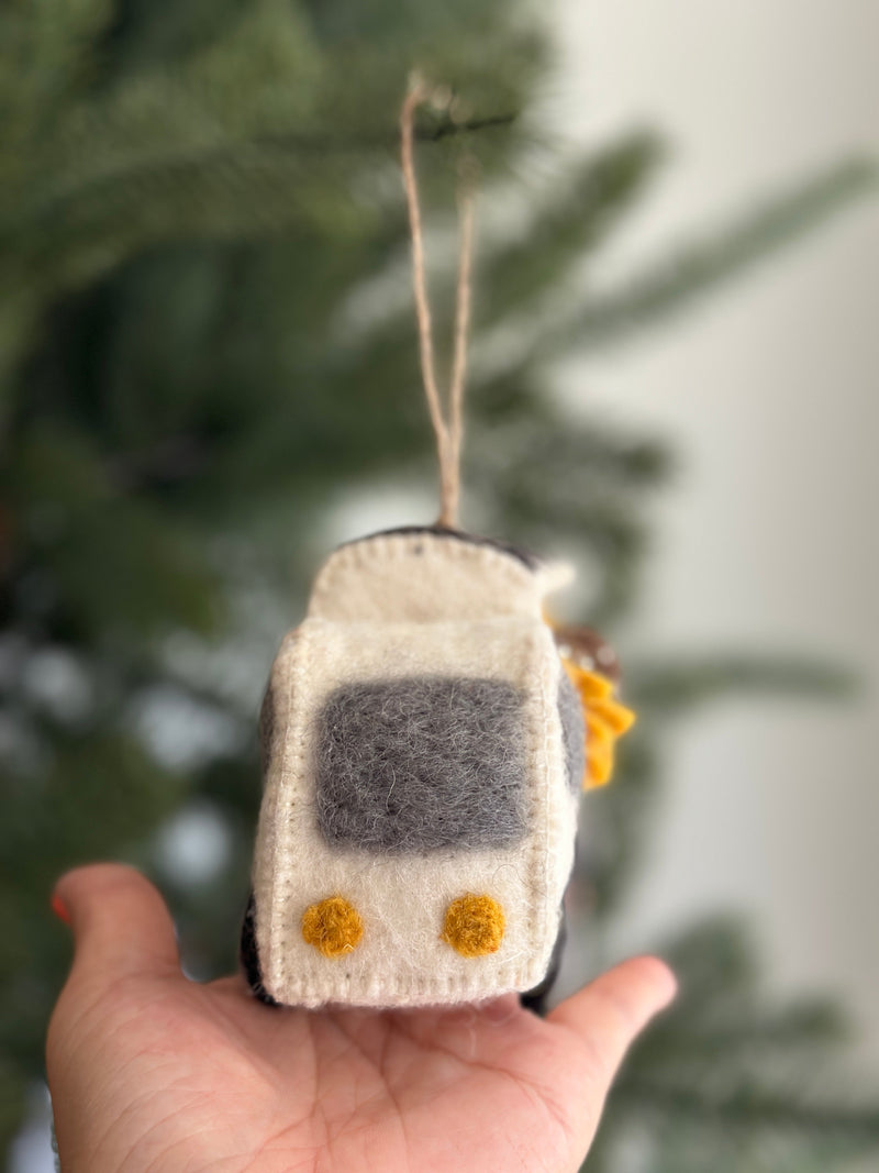 A handmade felt mobile flower shop Christmas ornament is displayed against a blurred Christmas tree background. This unique felt ornament features a white truck with a gray window, overflowing with vibrant sunflowers. A jute string is attached to the top for hanging.
