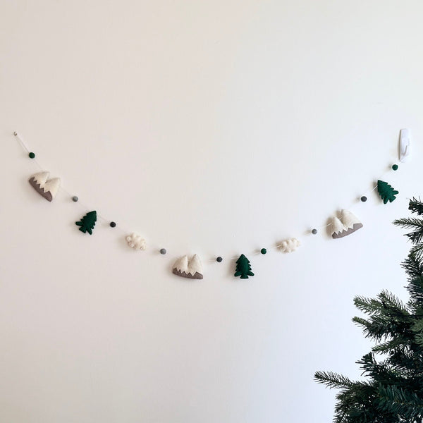 A playful wool felt mountain and cloud garland hangs against a bright white wall, featuring nature-inspired 3D designed felt figures. This unique garland showcases two-tone gray and white mountains, deep green trees, and white clouds, all connected by string and interspersed with felt balls. The handmade felt garland is secured by a transparent hook, with a green Christmas tree visible in the lower right.