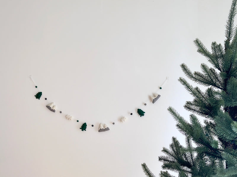 A wool felt mountain and cloud garland is displayed against a white wall, with a large pine tree partially visible on the right. The handcrafted garland features alternating 3D wool felt shapes of dark green pine trees, grey and white mountains, and fluffy white clouds. Small grey and dark green felt balls separate these elements along a white cord.