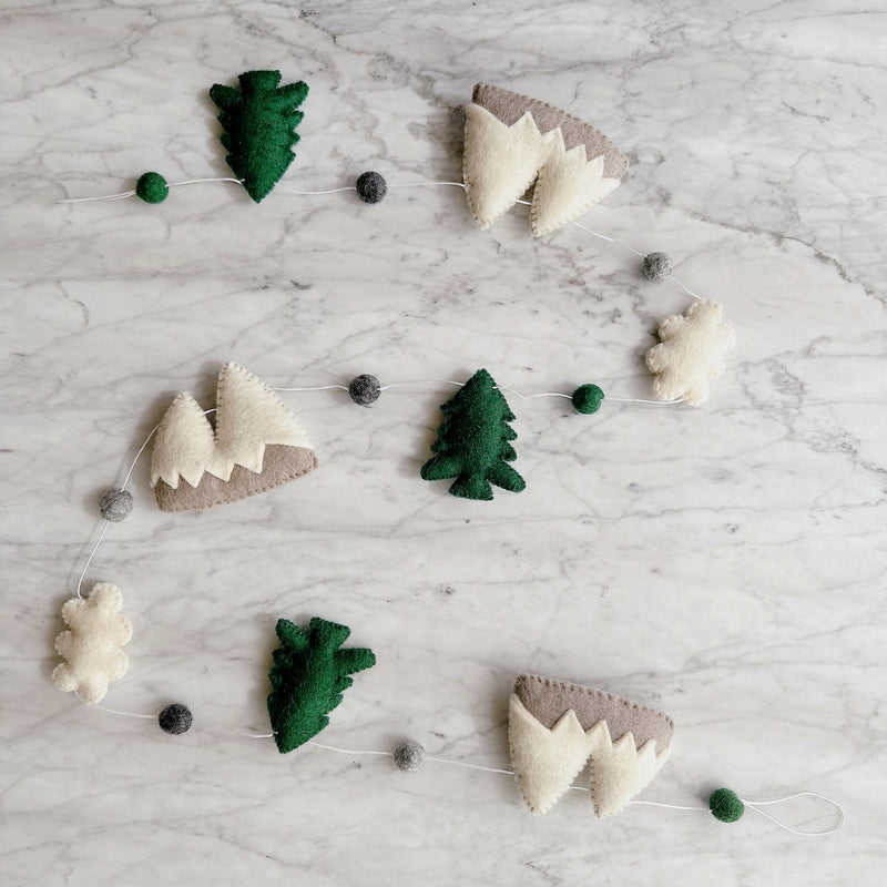 A wool felt Mountain & Cloud garland rests on a mottled white surface. This charming forest-themed garland showcases alternating 3D shapes of green trees, white clouds, and two-tone mountains. Small green and grey felt balls are interspersed, all connected by a thin white string.