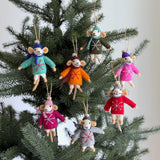 A set of seven wool felt mouse Christmas ornaments, each wearing a sweater, hangs on a festive Christmas tree, creating a joyful atmosphere. Each felt mouse ornament comes in a unique color like red, pink, orange, green, blue, purple, or gray, sporting an embroidered snowflake sweater with a matching hat and scarf. Crafted from soft wool felt, these charming mice are suspended by rustic twine loops.