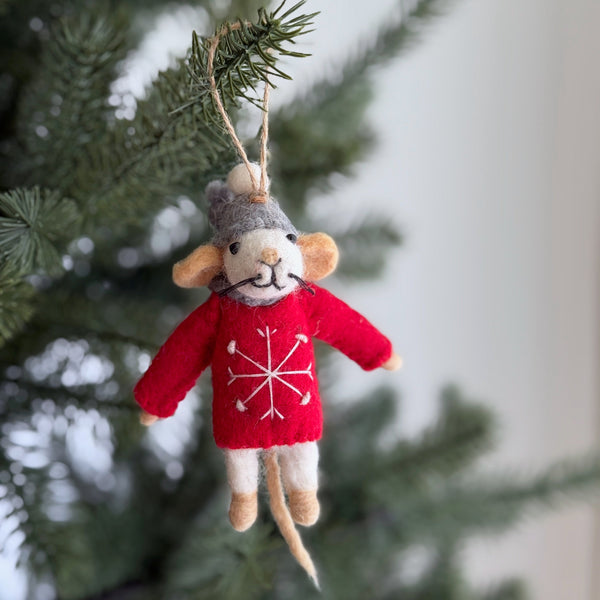 A handcrafted wool felt mouse Christmas ornament hangs on a festive pine branch. The felt mouse ornament wears a gray hat and a red sweater embroidered with a white snowflake. It showcases delicate ears, tiny black eyes, and a slender tail, all crafted from soft wool felt. A jute string is attached for hanging, adding a whimsical touch.