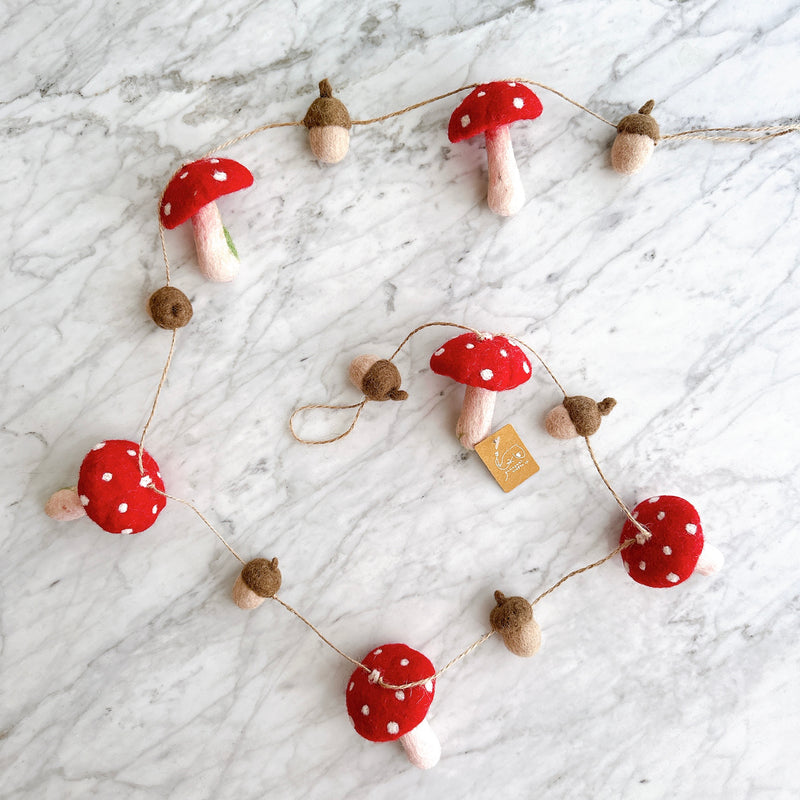 A whimsical felt mushroom and acorn garland rests on a white marble surface. This charming felt wool garland features alternating red mushrooms with white spots and tan acorns, strung along a thin twine. Its gentle curve creates an inviting display.
