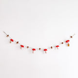 A cheerful felt mushroom and acorn garland is displayed on a plain white wall. This handcrafted wool felt garland features alternating red mushrooms with white dots on their caps and brown acorns, strung and secured by transparent hooks.