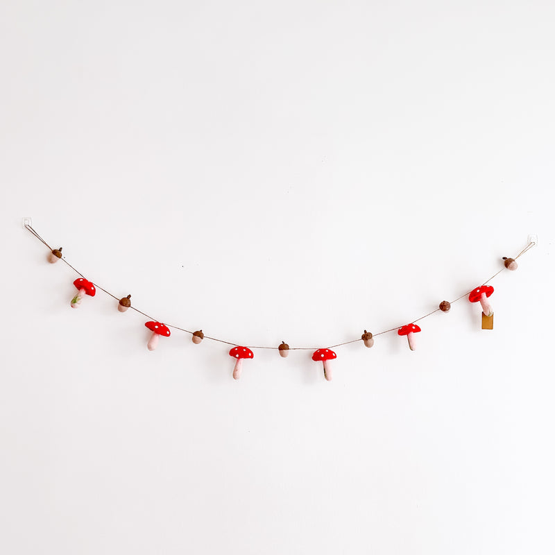 A cheerful felt mushroom and acorn garland is displayed on a plain white wall. This handcrafted wool felt garland features alternating red mushrooms with white dots on their caps and brown acorns, strung and secured by transparent hooks.