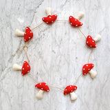 A felt mushroom garland is playfully arranged in a circular shape on a textured, light gray marble surface. The charming garland features multiple red mushroom caps with white spots and off-white stems, connected by thin, rustic twine, creating a whimsical and handcrafted appeal.