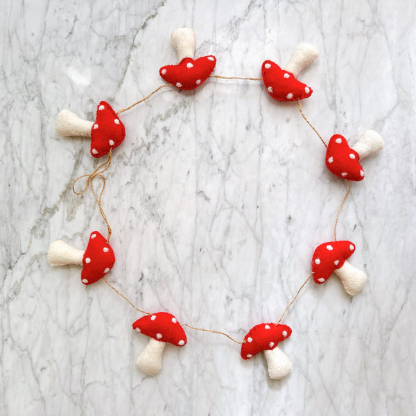 A felt mushroom garland is playfully arranged in a circular shape on a textured, light gray marble surface. The charming garland features multiple red mushroom caps with white spots and off-white stems, connected by thin, rustic twine, creating a whimsical and handcrafted appeal.