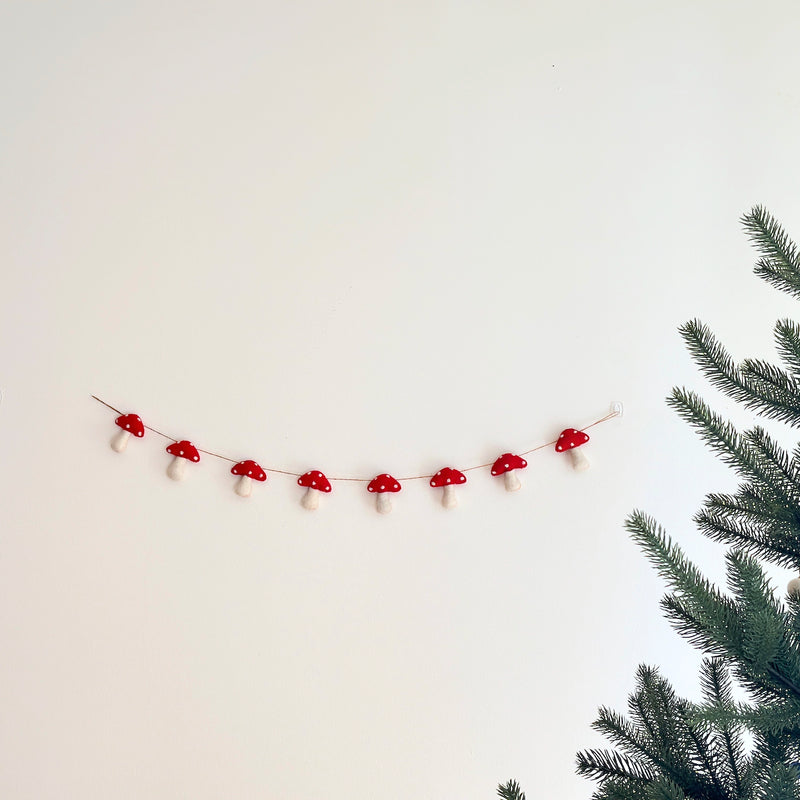A felt mushroom garland is displayed against a plain white wall next to a lush green pine tree branch. This charming felt garland features several evenly spaced felt mushrooms, each with a vibrant red cap dotted with white spots and a soft, white stem. The mushrooms are strung on a thin, light brown string, attached to the wall with clear fasteners, evoking a whimsical natural scene.