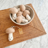 Multiple handmade felt mushroom keychains are charmingly displayed, some nestled in a neutral ceramic bowl and one resting on a woven brown mat, all against a clean white marble surface. Each whimsical felt mushroom keychain features a smiling face, a light brown cap, and a white stem, complete with a metal key ring. One of the unique keychains also includes a small tag attached with twine, adding to its handcrafted appeal.