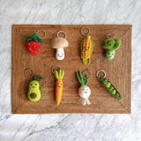 Eight charming, handmade felt keychains lay on a brown woven mat, set against a white and grey marble surface. Each keychain is a different smiling fruit or vegetable. In the top row, there is a red strawberry, a beige mushroom, a yellow corn on the cob, and a green broccoli floret. The bottom row features a green avocado half, an orange carrot, a white turnip, and a green pea pod with three peas peeking out. All have a silver metal keyring attached.