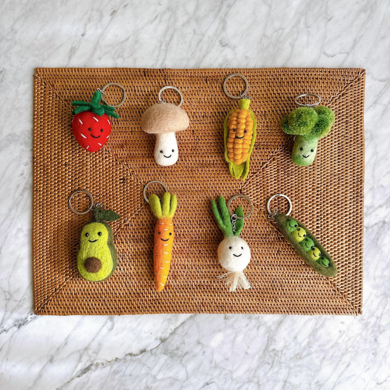 Eight charming, handmade felt keychains lay on a brown woven mat, set against a white and grey marble surface. Each keychain is a different smiling fruit or vegetable. In the top row, there is a red strawberry, a beige mushroom, a yellow corn on the cob, and a green broccoli floret. The bottom row features a green avocado half, an orange carrot, a white turnip, and a green pea pod with three peas peeking out. All have a silver metal keyring attached.