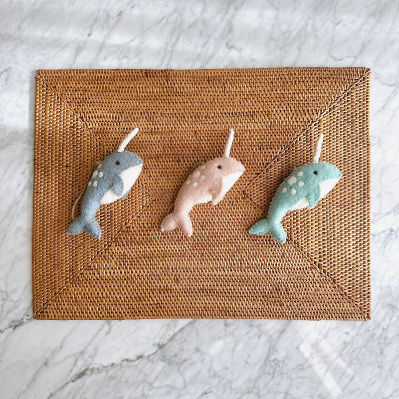 Three Felt Narwhal Ornaments are playfully displayed on a rectangular woven rattan placemat, set against a smooth marble backdrop. Each unique handcrafted felt narwhal features a distinct white horn and small white body dots, with jute strings for hanging. They come in charming light blue, pink, and green colors.