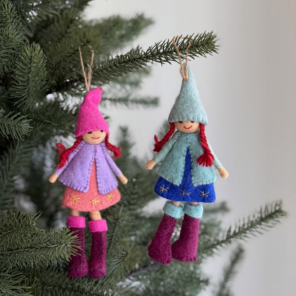 Two felt Nordic girl ornaments hang festively on a Christmas tree, each with distinct colors and textures. One ornament, dressed in a pink pointy hat, light purple coat, and pink star-embroidered dress with fuchsia boots, hangs beside another wearing a light blue hat, light blue coat, and dark blue star-embroidered dress with fuchsia boots. They are suspended by jute strings, adding a rustic, whimsical touch.