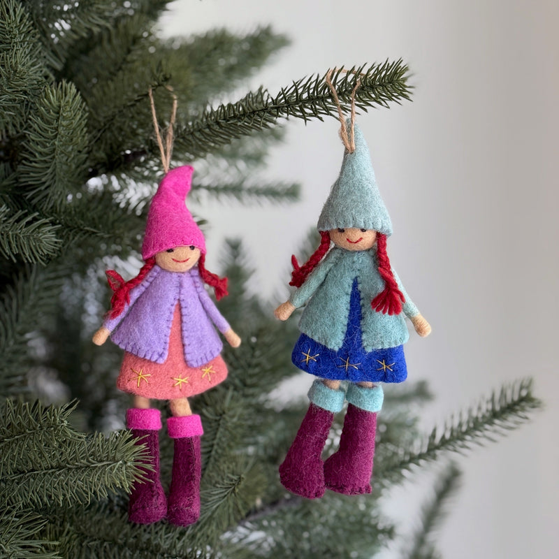 Two felt Nordic girl ornaments hang festively on a Christmas tree, each with distinct colors and textures. One ornament, dressed in a pink pointy hat, light purple coat, and pink star-embroidered dress with fuchsia boots, hangs beside another wearing a light blue hat, light blue coat, and dark blue star-embroidered dress with fuchsia boots. They are suspended by jute strings, adding a rustic, whimsical touch.