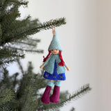 A charming wool felt Nordic girl ornament hangs from a lush green Christmas tree branch. The handcrafted girl features a light blue pointed hat, long red braids, and a cheerful smile. She wears a light blue wool felt coat over a dark blue skirt with yellow star details, complemented by maroon boots.