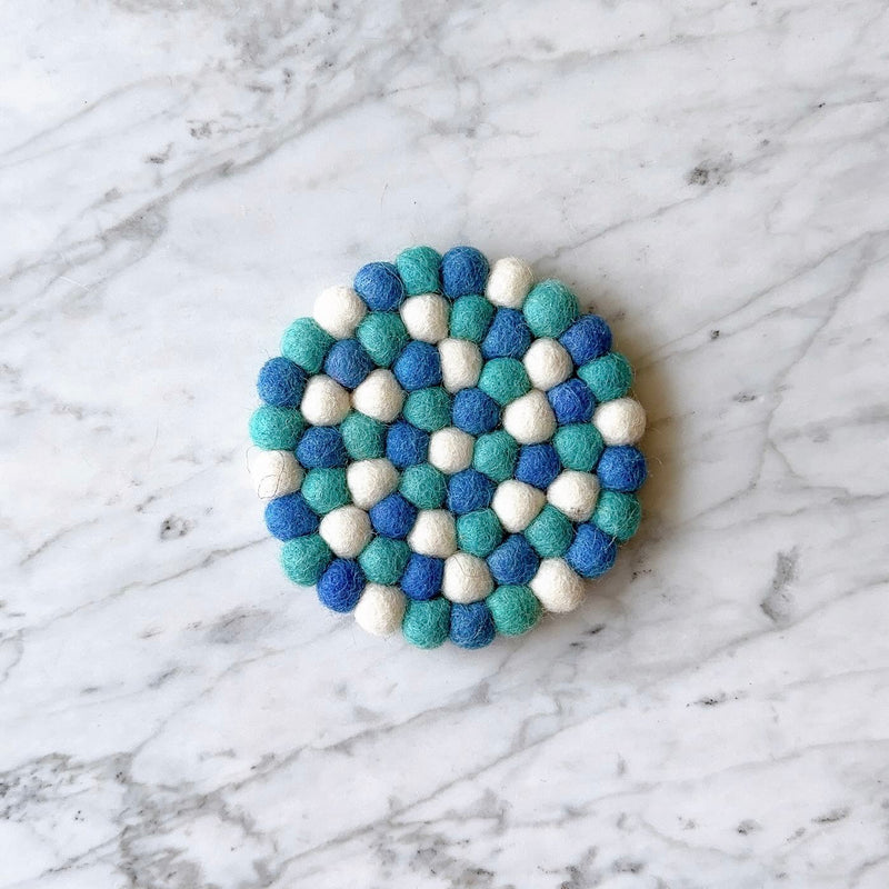 A handcrafted Ocean Blue Round felt coaster, made from 100% organic lamb wool, rests elegantly on a cool marble surface. This round wool felt coaster, approximately 4 inches in diameter, features a unique, tactile design of hand-stitched felt balls in alternating shades of ocean blue, white, and turquoise. Its serene aesthetic highlights the artisanal quality of this fair-trade piece, ethically made by Nepalese female artisans.