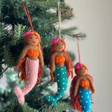 Three assorted handmade wool felt mermaid ornaments hang on a festive green Christmas tree. Each mermaid features an orange top, a pink flower in her hair, and a tail adorned with white dots. One has a pink tail, another a teal tail, and the third a blue tail, all with bendable arms and tails for posing.