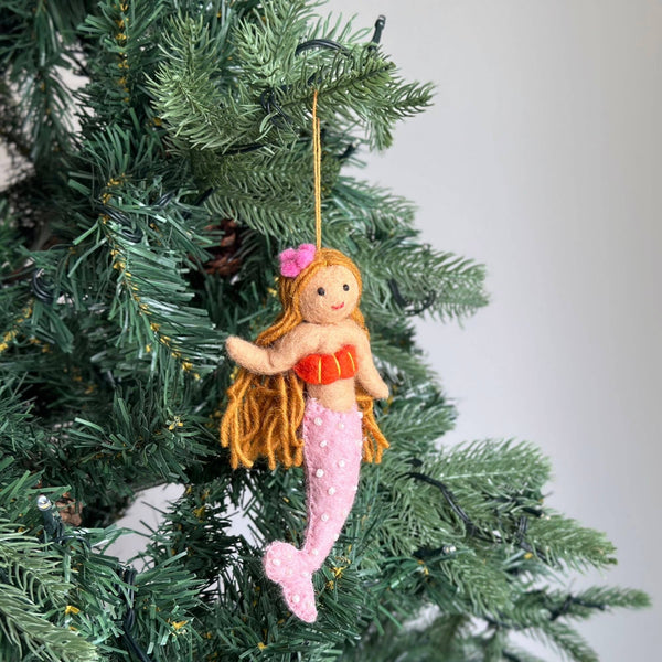 A handcrafted wool felt mermaid ornament hangs from a Christmas tree branch, held by a cotton string. This charming felt mermaid features golden yarn hair with a small pink flower, a red top, and a pink tail adorned with white dots. Its arms and tail are bendable for posing. The cheerful ornament is beautifully displayed against the dense green needles of the tree, creating a whimsical scene.