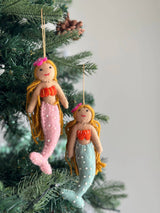 Two assorted wool felt mermaid ornaments hang from brown jute strings on a Christmas tree, set against a soft grey background. These handmade felt mermaids feature tan faces, stitched eyes, red smiles, and long golden yarn hair adorned with unique felt flowers. One has a pink beaded tail and orange seashell top, while the other has a light blue beaded tail and orange seashell top. Both have bendable arms and tails for posing.