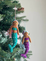 Two assorted handmade wool felt mermaid ornaments hang gracefully on green Christmas tree branches. One felt mermaid ornament has light brown skin, pink hair with a teal bow, and a teal tail with white dots, suspended by a pink string. The second felt mermaid ornament features light brown skin, blue hair with a purple bow, and a purple tail with white dots, hanging from a blue string, creating a whimsical and festive display.