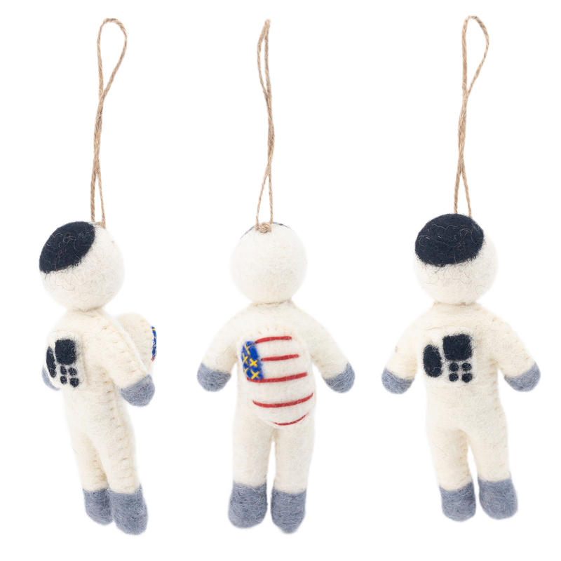 A handmade felt astronaut ornament is displayed against a bright white background, shown from three distinct angles: side, front, and back. This needle felted ornament features a white body with grey extremities and a black rounded square on the head. A flag graphic appears on the front chest and right arm, while the back displays a black square design.