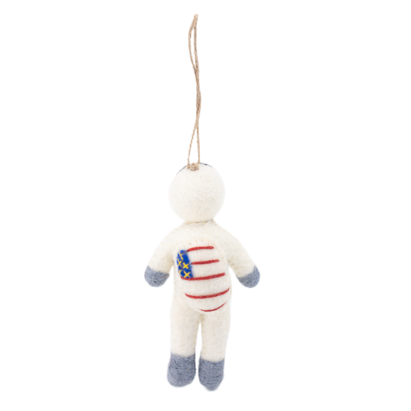 A whimsical felt astronaut ornament floats against a bright white background, suspended by a jute string. This handmade wool felt astronaut has a creamy white body, a spherical head, and soft gray felt tips on its arms and legs. Its chest features a stylized design of blue stars and red stripes, mimicking a flag, adding a sense of childlike wonder.