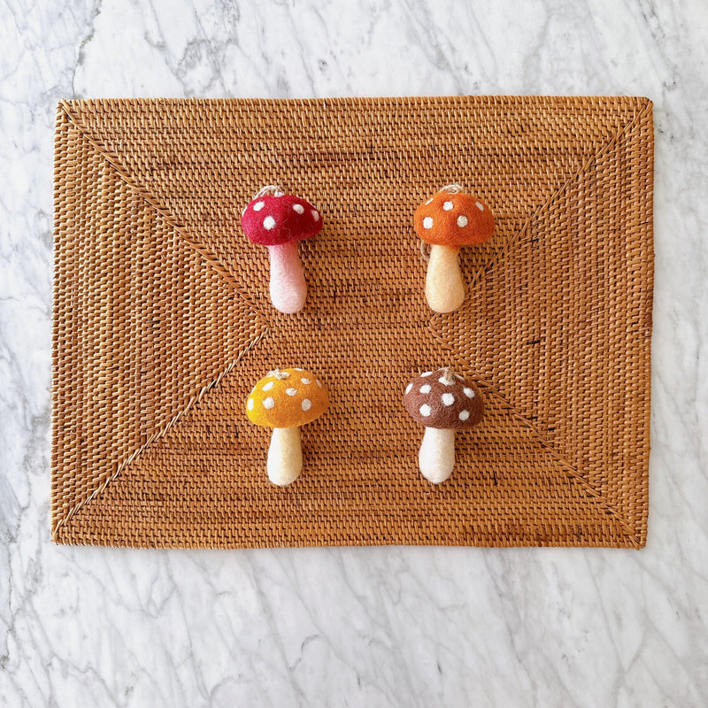 Autumn Tone Mushroom Set of 4 felt ornaments rest on a textured woven mat against a bright marble surface. Each handcrafted wool felt mushroom ornament features a charming toadstool design with a rounded, white polka-dotted cap in warm red, orange, yellow, or brown, and a cylindrical stem. A jute string tops each unique Christmas ornament, creating a natural, inviting display.