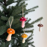A set of four autumn tone wool felt mushroom ornaments are displayed hanging on the branches of a Christmas tree, against a soft, neutral background. Each handmade felt mushroom ornament features a classic toadstool design with rounded caps in red, orange, yellow, or brown, adorned with white polka dots. Their soft off-white stems and thin jute strings add a rustic, whimsical touch, perfect for holiday decor.