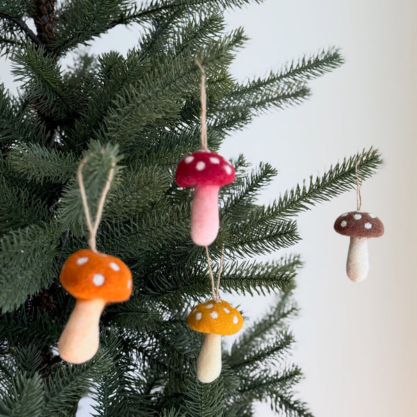 A set of four autumn tone wool felt mushroom ornaments are displayed hanging on the branches of a Christmas tree, against a soft, neutral background. Each handmade felt mushroom ornament features a classic toadstool design with rounded caps in red, orange, yellow, or brown, adorned with white polka dots. Their soft off-white stems and thin jute strings add a rustic, whimsical touch, perfect for holiday decor.