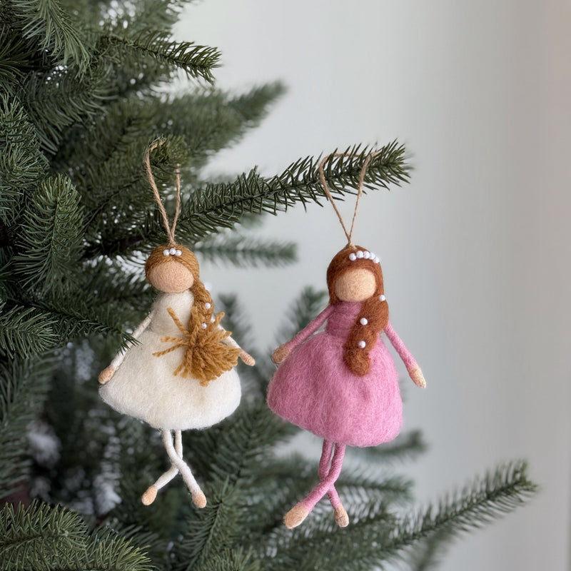 Two handcrafted wool felt ballerina ornaments hang gracefully from a lush Christmas tree. One ballerina wears a white dress with golden braided hair, adorned with small white beads. The other features a pink dress, brown hair, and also white beads. Both delicate figures have soft felt limbs and are suspended by jute strings, adding a whimsical touch.